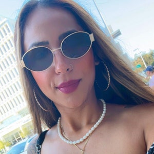 Puede incluir: Una mujer con el pelo largo y casta&ntilde;o lleva gafas de sol ovaladas con montura dorada y un collar de perlas. Lleva un aro de nariz dorado.