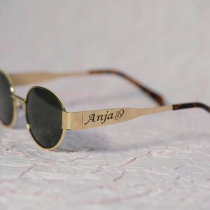Puede incluir: Gafas de sol redondas con montura dorada y lentes oscuras. La barra de metal en la montura est&aacute; grabada con el nombre "Anja" y un s&iacute;mbolo de coraz&oacute;n.