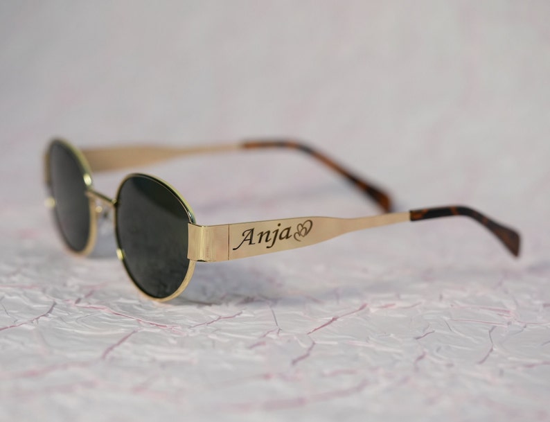 Puede incluir: Gafas de sol redondas con montura dorada y lentes oscuras. La barra de metal en la montura est&aacute; grabada con el nombre "Anja" y un s&iacute;mbolo de coraz&oacute;n.