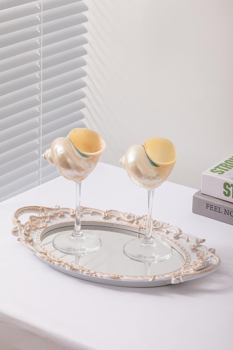 May include: Two seashell-shaped wine glasses with clear stems sit on a mirrored tray with an ornate, cream-coloured frame. The glasses have a pearlescent finish and a yellow interior. The tray rests on a white surface.