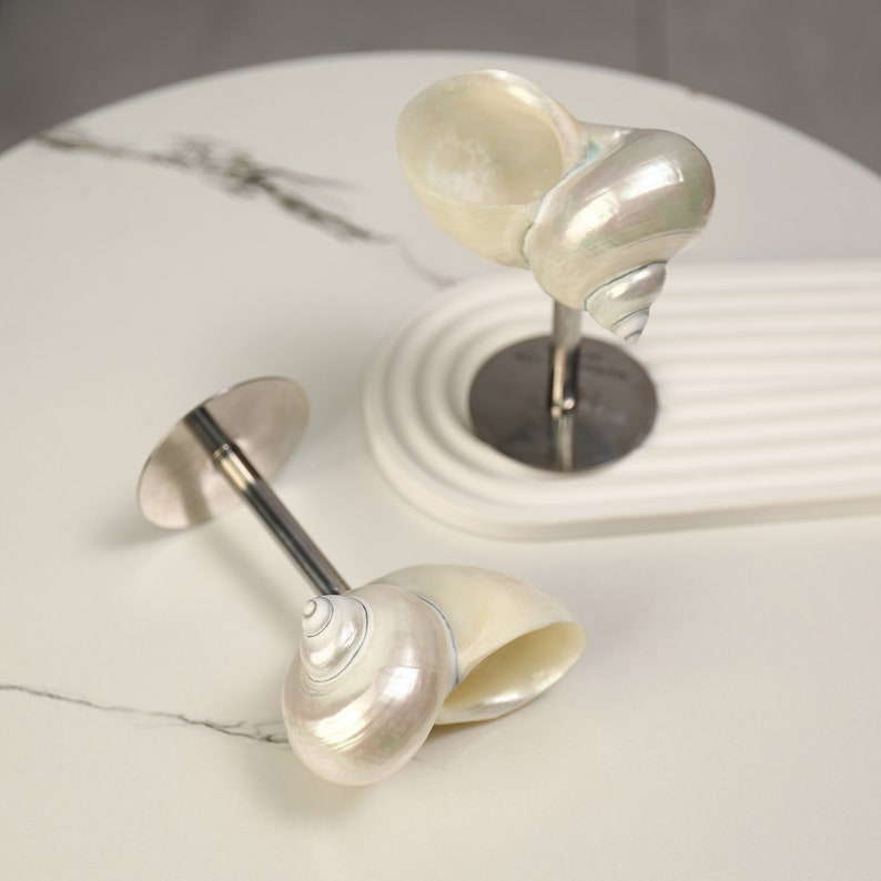 May include: Two decorative seashell sculptures. Each sculpture features a pearlescent nautilus shell mounted on a silver-coloured metal stem and base. One sculpture is upright, while the other is lying on its side. The shells have a natural, iridescent sheen.