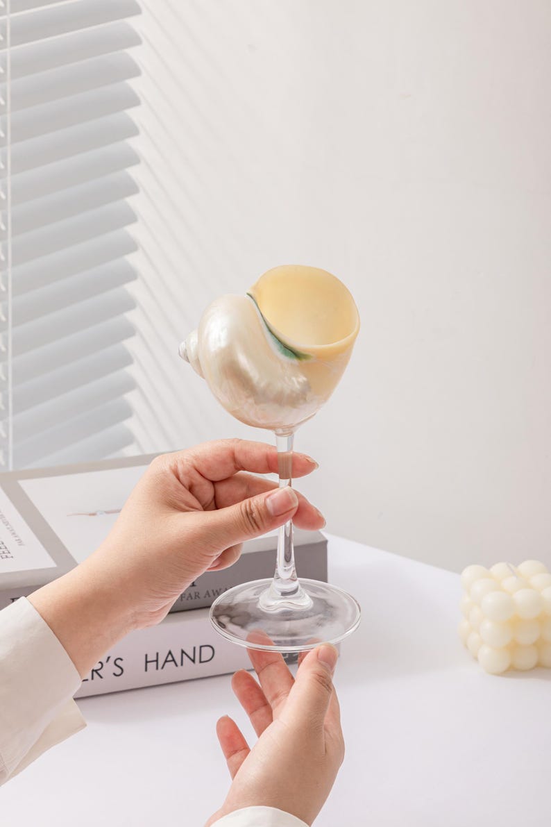 May include: A clear glass wine glass with a seashell-shaped bowl. The seashell is pearlescent white and yellow. The glass is held by a person's hands. The base of the glass is round.