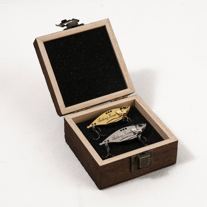 May include: A wooden box with a hinged lid, containing two fishing lures. The lures are silver and gold, and both have the name "Anthony Kivon" engraved on them.