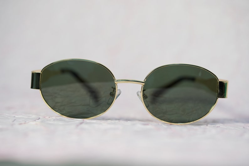 Puede incluir: Un par de gafas de sol redondas con montura dorada y lentes verde oscuro.