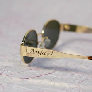 Puede incluir: Gafas de sol redondas con montura dorada y lentes oscuras. Las gafas tienen una banda de metal dorado con el nombre "Anja" y dos corazones grabados en el lateral.
