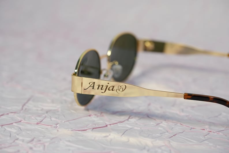 Puede incluir: Gafas de sol redondas con montura dorada y lentes oscuras. Las gafas tienen una banda de metal dorado con el nombre "Anja" y dos corazones grabados en el lateral.
