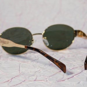 Puede incluir: Gafas de sol ovaladas con montura dorada y patillas de carey. Las gafas tienen lentes verdes y la patilla izquierda tiene la palabra "Anja" grabada.