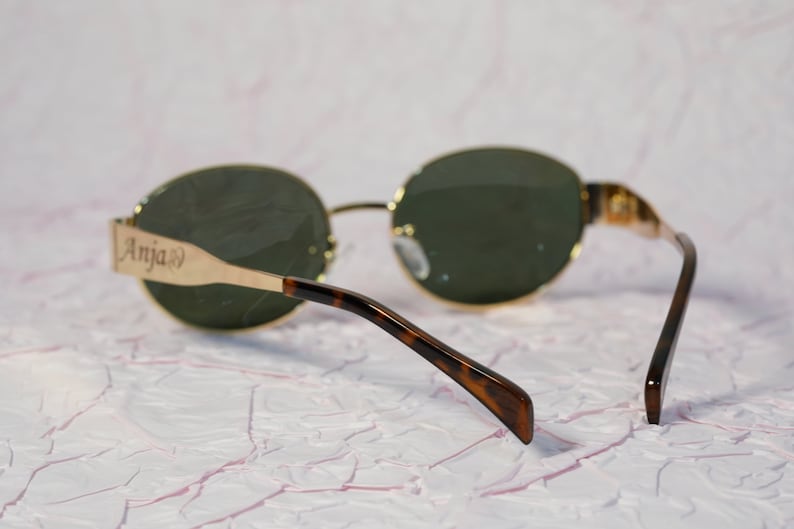 Puede incluir: Gafas de sol ovaladas con montura dorada y patillas de carey. Las gafas tienen lentes verdes y la patilla izquierda tiene la palabra "Anja" grabada.