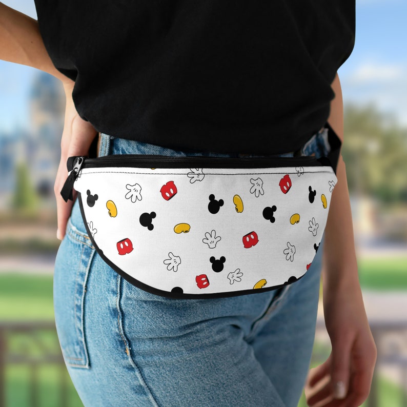 Puede incluir: Ri&ntilde;onera blanca con cremallera y ribete negros. La ri&ntilde;onera tiene un estampado de cabezas de Mickey Mouse negras, jud&iacute;as amarillas, pantalones cortos rojos y guantes blancos. Se lleva en la cintura.