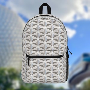 May include: A backpack with a geometric pattern in shades of gray, white, and light blue. The backpack has a black zipper, a front pocket, and a mesh side pocket. The background is blurred, showing buildings and a blue sky.