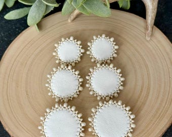 May include: Six white leather earrings with a pearl-like beaded border are arranged on a wooden surface. The earrings are circular, with a stitched edge. Green foliage and a deer antler are in the background.