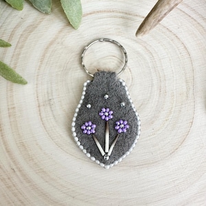 May include: A grey felt keyring with a floral design. The keyring features three purple beaded flowers with white accents and a silver key ring. The edges are lined with small white beads.
