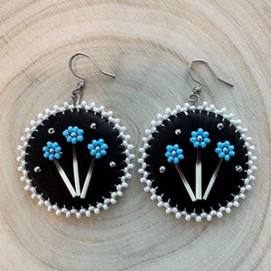 May include: Handmade black leather earrings with a white beaded edge. Each earring features three light blue beaded flowers with white quill accents and silver-coloured studs. The earrings have silver-coloured ear wires.