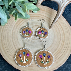 Peut inclure: Une paire de boucles d'oreilles en perles avec un motif floral. Les boucles d'oreilles sont en cuir brun et ont des perles blanches autour des bords. Les fleurs sont violettes, roses et bleues.