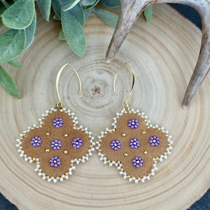 May include: A pair of gold dangle earrings with a brown suede and purple flower design. The earrings are surrounded by white beads.