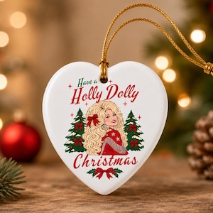 May include: A white heart-shaped ceramic ornament featuring a graphic illustration of a blonde woman with voluminous hair wearing a red outfit, flanked by two green Christmas trees, with the text Have a Holly Dolly Christmas in red and green.