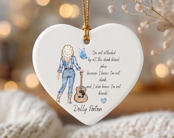 May include: A white heart-shaped ceramic ornament hanging from a gold cord. It features an illustration of a blonde person in denim holding a guitar, a blue butterfly, a small blue heart, and the text: I'm not offended by all the dumb blonde jokes because I know I'm not dumb... and I also know I'm not blonde. Dolly Parton.