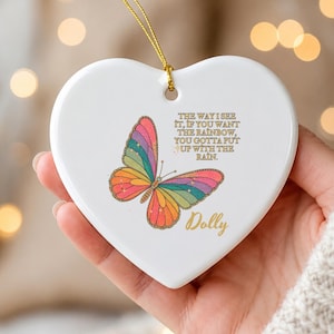May include: A white heart-shaped ceramic ornament with a gold string. It features a colorful rainbow butterfly graphic and the text: THE WAY I SEE IT, IF YOU WANT THE RAINBOW, YOU GOTTA PUT UP WITH THE RAIN. The name Dolly is printed below.