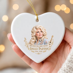 May include: A white heart-shaped ceramic ornament featuring a portrait of Dolly Parton with a guitar. The text reads: HER MUSIC WILL SHINE FOREVER, DOLLY PARTON, 1946-2026. It includes a gold hanging string and decorative gold musical notes.