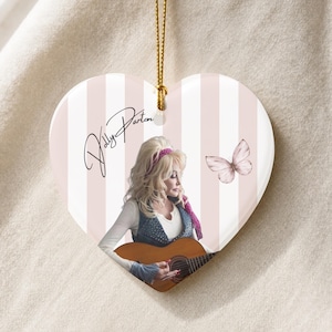 May include: A heart-shaped hanging ornament featuring a light pink and white vertical striped background. The design includes a portrait of Dolly Parton playing an acoustic guitar, a butterfly illustration, and the signature Dolly Parton in black script.