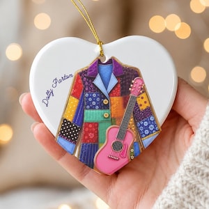 May include: A heart-shaped white ceramic ornament featuring a colorful patchwork jacket illustration and a pink acoustic guitar. The text Dolly Parton is printed in blue script on the left side. A gold string is attached to the top for hanging.