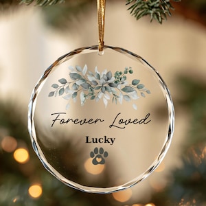 May include: A circular clear glass ornament with faceted edges, featuring a printed design of green eucalyptus leaves, the text Forever Loved, the name Lucky, and a dark gray paw print icon with a small heart in the center, suspended by a gold cord.