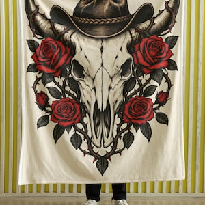 May include: A cream-colored throw blanket featuring a central graphic of a white bull skull wearing a brown cowboy hat. The skull is surrounded by vibrant red roses and dark green leaves intertwined with thorny brown vines.