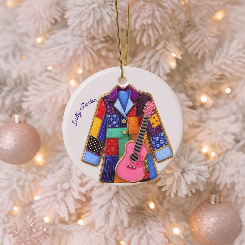 May include: A round white ceramic ornament featuring a colorful patchwork coat illustration with a pink acoustic guitar overlaid on the front. The text "Dolly Parton" is printed in blue script on the left side. A gold string is attached to the top.