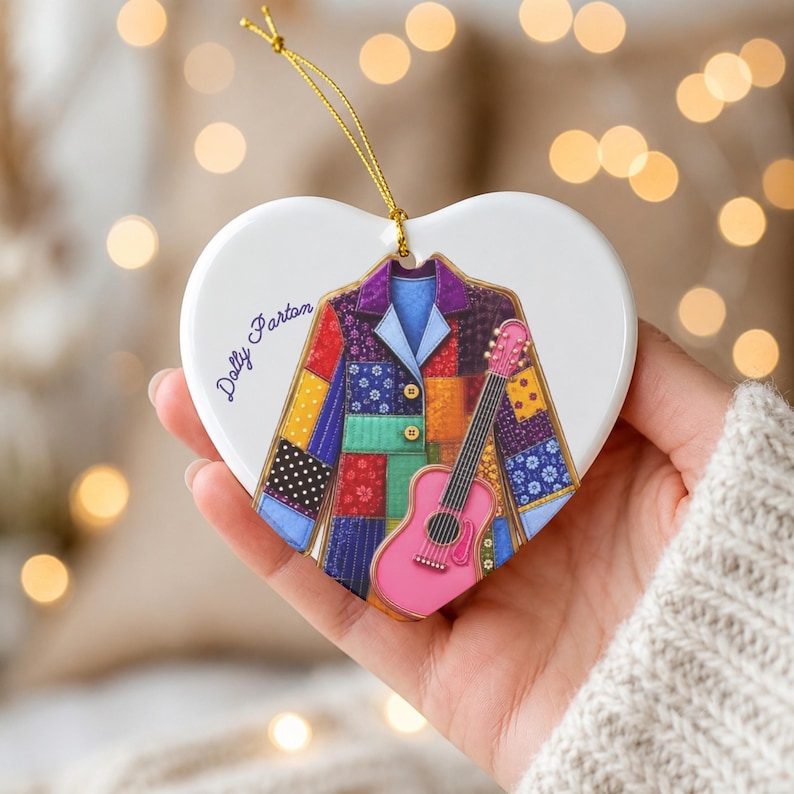 May include: A white heart-shaped ceramic ornament featuring a colorful patchwork jacket illustration and a pink acoustic guitar. The text Dolly Parton is printed in black script on the left side. A gold string is attached to the top for hanging.