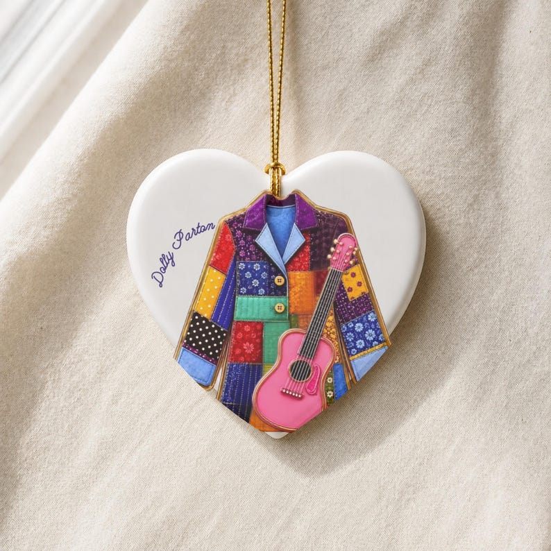 May include: A white heart-shaped ceramic ornament hanging from a gold string. The center features a colorful patchwork jacket illustration with a pink acoustic guitar and the text Dolly Parton written in blue script on the left side.