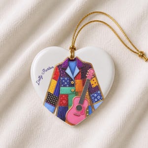 May include: A heart-shaped white ceramic ornament featuring a colorful patchwork jacket illustration and a pink acoustic guitar. The text Dolly Parton is printed in blue script on the left side. A gold string is attached to the top for hanging.