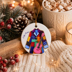 May include: A round white ceramic ornament featuring a colorful patchwork coat illustration and a pink acoustic guitar. The text Dolly Parton is printed in blue on the left side. The ornament hangs from a thin gold string for display purposes.