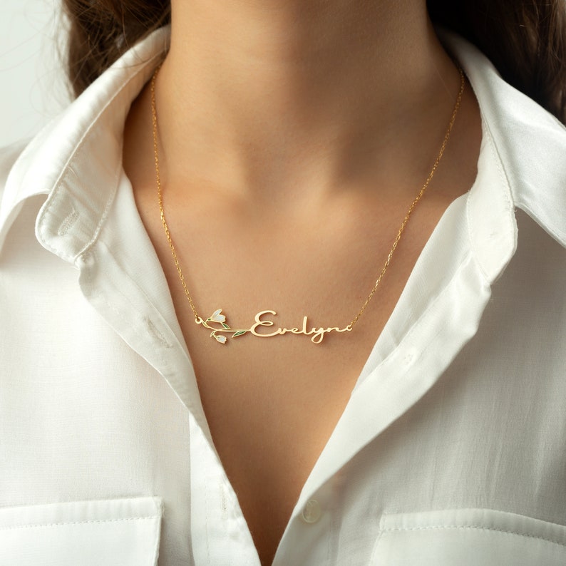 May include: A gold necklace with the name "Evelyn" written in cursive script with a delicate floral design. The necklace is on a model wearing a white shirt.