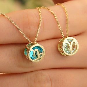 May include: Two gold-colored necklaces with round pendants. Each pendant features a gemstone and a zodiac symbol. One pendant has a blue gemstone with a Capricorn symbol, and the other has a green gemstone with an Aries symbol.