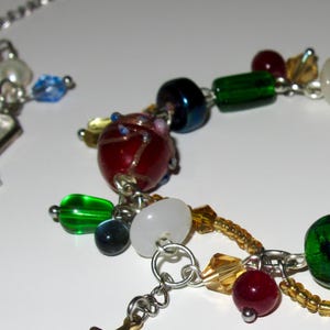 Op de afbeelding: Een kleurrijke armband met kralen en diverse bedels. De armband heeft rode, groene, witte en blauwe kralen, evenals een gouden sterbedel. De armband is gemaakt van zilverkleurig metaal.