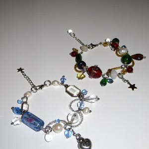 Op de afbeelding: Twee bedelarmbanden met kleurrijke kralen. E&eacute;n armband heeft rode, groene en gele kralen. De andere heeft blauwe, witte en transparante kralen, met een hartbedel en een sterbedel. De armbanden zijn van zilverkleurig metaal.