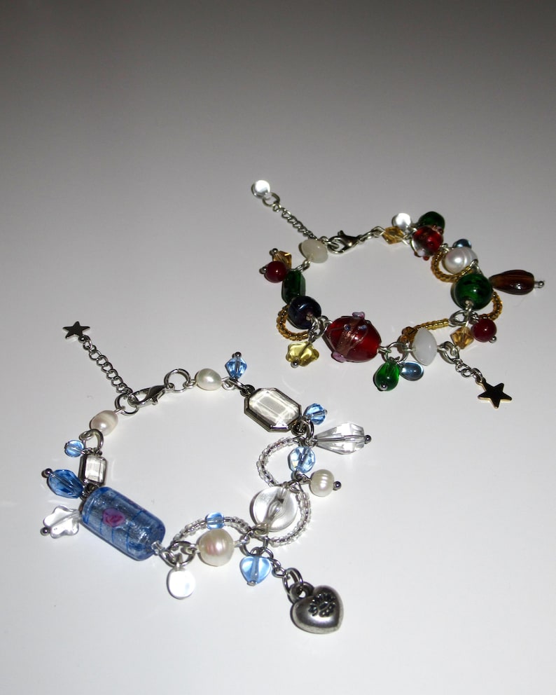 Op de afbeelding: Twee bedelarmbanden met kleurrijke kralen. E&eacute;n armband heeft rode, groene en gele kralen. De andere heeft blauwe, witte en transparante kralen, met een hartbedel en een sterbedel. De armbanden zijn van zilverkleurig metaal.