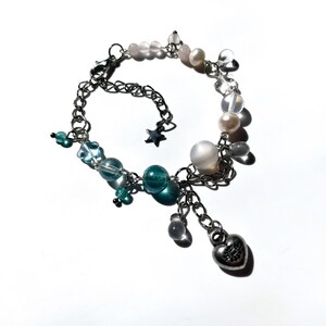 Puede incluir: Una pulsera de cadena plateada con una variedad de cuentas y dijes. La pulsera presenta cuentas transparentes, azules y rosas, junto con perlas y un dije en forma de coraz&oacute;n. Tambi&eacute;n se incluye un peque&ntilde;o dije de estrella.
