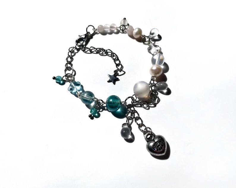 Puede incluir: Una pulsera de cadena plateada con una variedad de cuentas y dijes. La pulsera presenta cuentas transparentes, azules y rosas, junto con perlas y un dije en forma de coraz&oacute;n. Tambi&eacute;n se incluye un peque&ntilde;o dije de estrella.