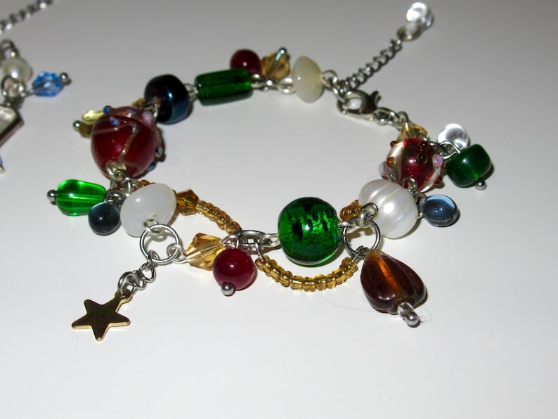 Op de afbeelding: Een kleurrijke armband met kralen en diverse bedels. De armband heeft rode, groene, witte en blauwe kralen, evenals een gouden sterbedel. De armband is gemaakt van zilverkleurig metaal.