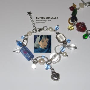 Op de afbeelding: Een armband met een zilveren ketting, versierd met blauwe, witte en transparante kralen, een hartvormige bedel en een kleine afbeelding van een anime-personage. De tekst "SOPHIE BRACELET" is zichtbaar.
