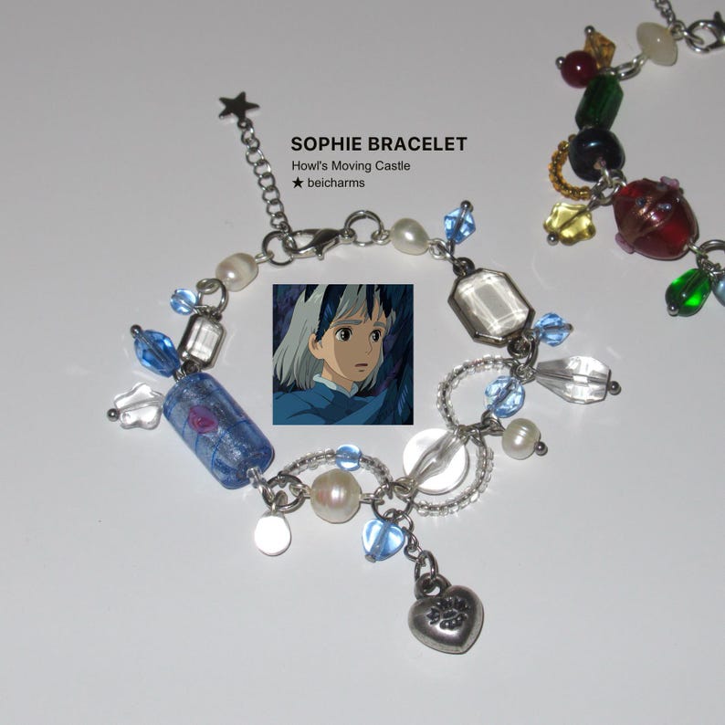 Op de afbeelding: Een armband met een zilveren ketting, versierd met blauwe, witte en transparante kralen, een hartvormige bedel en een kleine afbeelding van een anime-personage. De tekst "SOPHIE BRACELET" is zichtbaar.