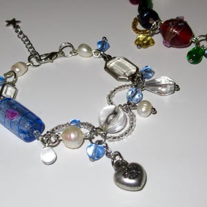 Op de afbeelding: Een zilveren armband met diverse bedels, waaronder blauwe en heldere kralen, parels en een hartvormige hanger. De armband heeft ook een blauw-paarse cilindrische kraal en een stervormige bedel.