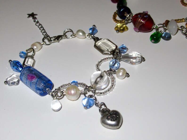 Op de afbeelding: Een zilveren armband met diverse bedels, waaronder blauwe en heldere kralen, parels en een hartvormige hanger. De armband heeft ook een blauw-paarse cilindrische kraal en een stervormige bedel.