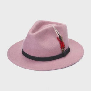 Peut inclure: Un chapeau fedora rose poudr&eacute; avec un bandeau noir et une plume d&eacute;corative. Le chapeau a un bord classique et une couronne pinc&eacute;e. La plume est rouge, noire et blanche.
