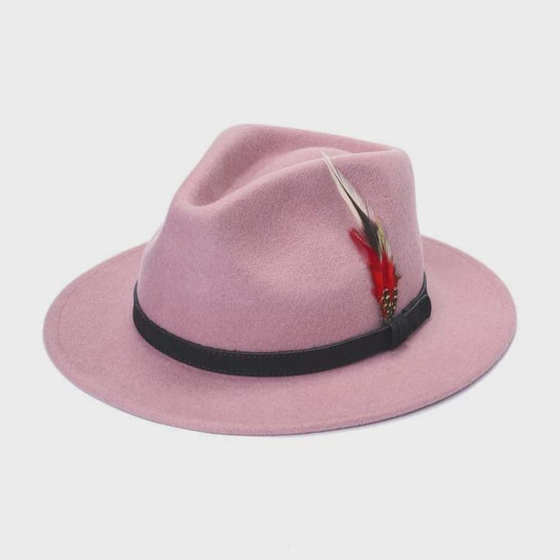 Peut inclure: Un chapeau fedora rose poudr&eacute; avec un bandeau noir et une plume d&eacute;corative. Le chapeau a un bord classique et une couronne pinc&eacute;e. La plume est rouge, noire et blanche.
