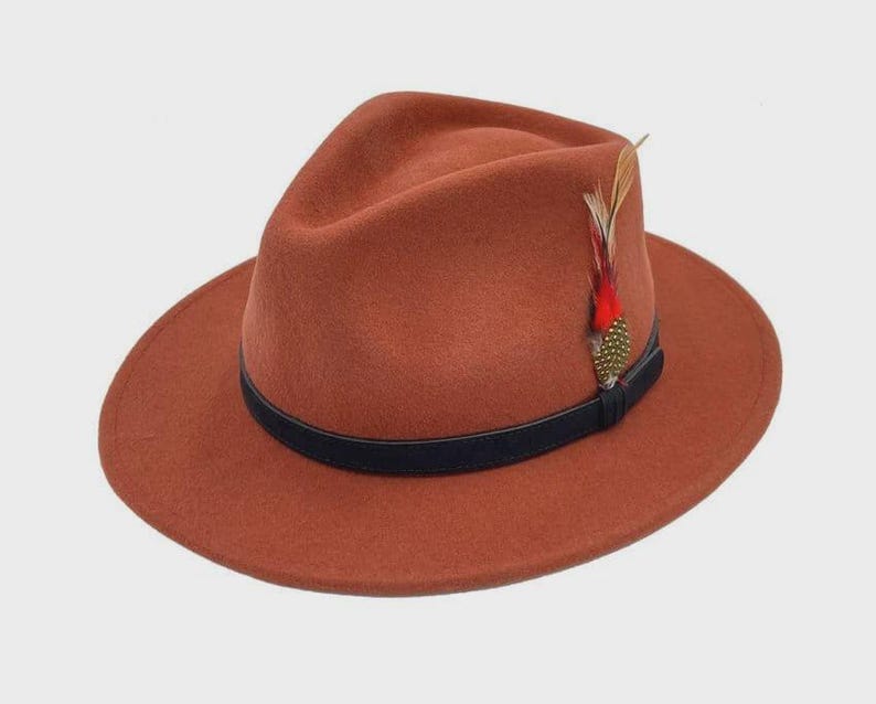 Peut inclure: Un chapeau fedora de couleur rouille avec une bande noire et une plume d&eacute;corative. Le chapeau a un large bord et un design classique. La plume est rouge, dor&eacute;e et verte.
