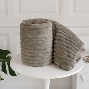 May include: A rolled-up, soft, grey blanket with a ribbed texture. The blanket is displayed on a white, round table. The blanket's plush material is visible, suggesting comfort and warmth. The background features a white wall and a green plant.