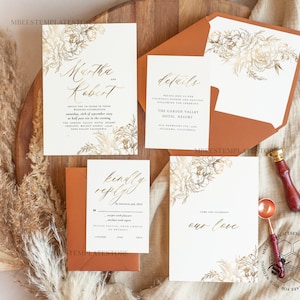 Puede incluir: Un conjunto de tarjetas de invitación de boda con diseños florales dorados y texto en caligrafía. Las tarjetas están sobre una superficie de madera marrón con hierba de la pampa seca.