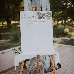 Op de afbeelding: Een wit bord met de tekst "he or she" en "WHAT WILL BABY SMITH BE?"  Write your name to cast your vote.  boy  girl" op een houten ezel.